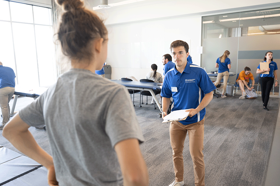 A male student with a blue polo shirt standing in the middle of a large examing room and talking to a clent with her back turned to the camera. 