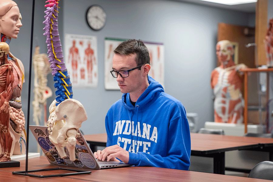 A man with short, dark brown hair and glasses, types on his computer in an anatomy lab. He wears a blue Indiana State hoodie.  There are spine and human body models around him in the lab.