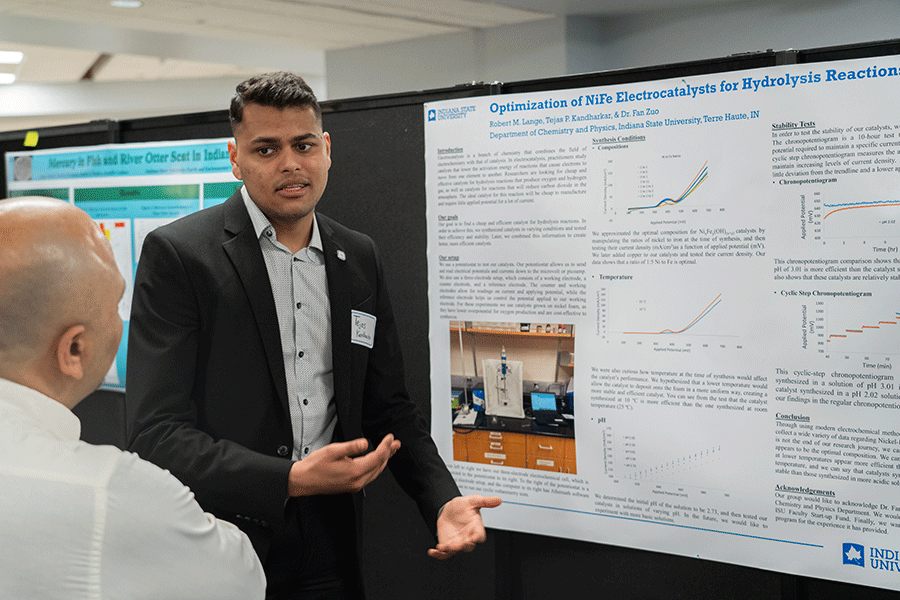 A male student of color with short black hair, wearing a black suit jacket and gray, open-collared shirt, presents his Honors research poster to an onlooker, an older man with a bald head and wearing a white shirt. The poster depicts photos, charts, and graphs related to the student’s research. Another blue, green, and white poster is visible in the background.   