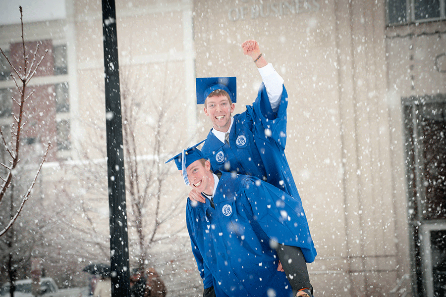 Two white male students in blue commencement cap and gown celebrate their graduation during a heavy winter snowfall outside of Federal Hall