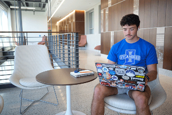  male student with a blue Indiana State University t-shirt works on a laptop in the lobby of a building 