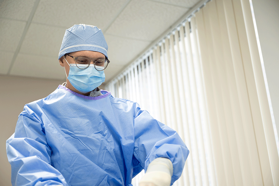 A white male physician wearing a blue surgical gown, a blue surgical mask, white surgical gloves, and a blue surgical cap. He also has dark brown glasses. He is looking down at his hands in a room with windows and window blinds visible behind him.   