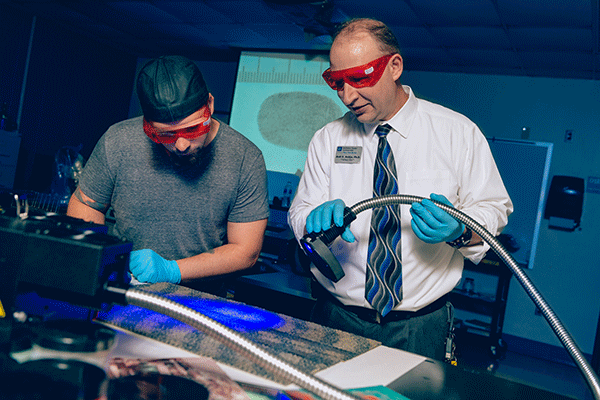 A male criminology professor with a white shirt and tie and red safety glasses shines a blue light on a sample as a student looks on with a projection of a fingerprint in the background.