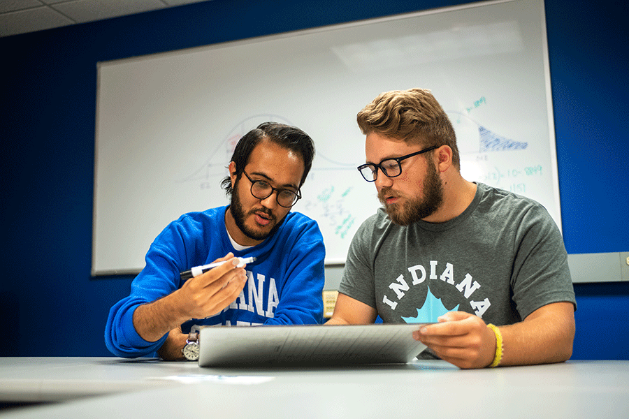 Two men sitting together in a classroom look like they are solving a problem on the white erase board in front of them. The man on the left has an olive complexion, black hair, beard, and moustache, and wears glasses. He is wearing an Indiana State sweatshirt with the sleeves rolled up and holds a black dry-erase marker in his hand. 