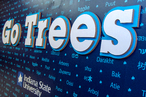 A navy-blue wall with a collage of words written in light blue ink on it. The words are in a variety of languages. The words include “Darakht,” “Puut,” “Arbores,” and “Bome.” “Go Trees” is on the wall in large white and blue lettering. The Indiana State University logo is below.
