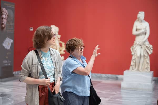 Two individuals are standing inside an art gallery with a red wall in the background.