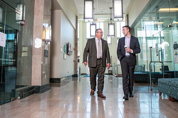 A WHITE PROFESSOR AND WHITE STUDENT, BOTH IN PROFESSIONAL ATTIRE, WALKING TOWARDS THE CAMERA AND TALKING IN A BRIGHTLY LIT HALLWAY. 