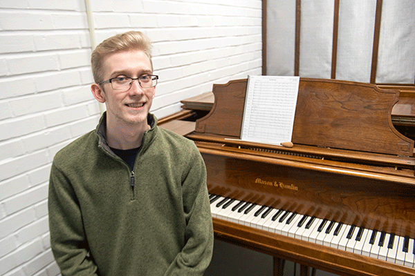 A white male student with blond hair and glasses sits on a piano bench in front of a brown piano. A book of sheet music sits atop the piano. There is a white brick wall in the background.