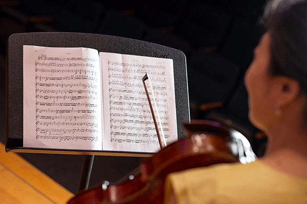 A book of sheet music is open on a black music stand. Blurred in the foreground, a woman with short black hair wearing a white blouse is holding a violin on her shoulder, with the bow extending up from her other hand. The background of the image is very dark.