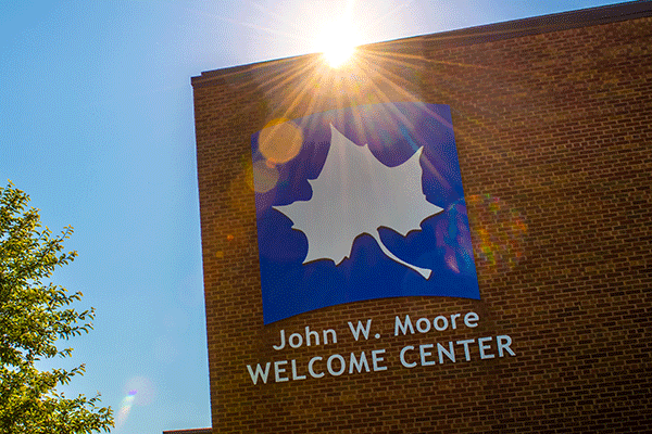 A brick building on the right of the image with a large Indiana State logo and “Welcome Center” text with the sun peeking over the building. A tree is visible in the left side of the frame with a blue sky.