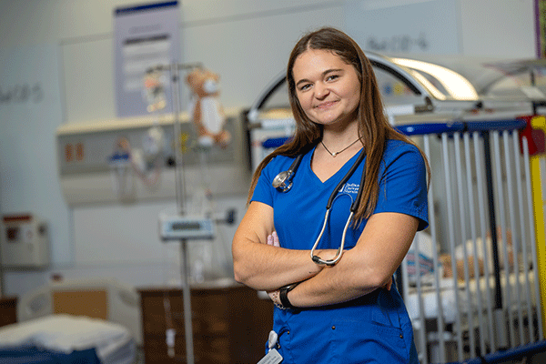 A female nursing student poses for the camera with a nursing lab in the background.