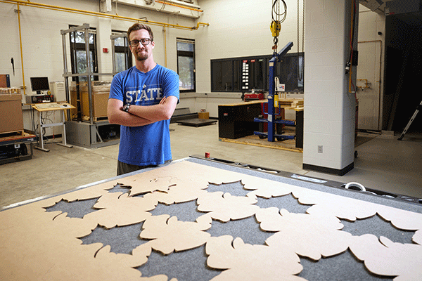 A picture of a white male student with his arms crossed, standing behind a large piece of cardboard on a worktable that has Sycamore leaf shapes cut out of it. The man has dark brown hair, a beard, and a moustache, and wears glasses. He is wearing a blue t-shirt that says STATE and is standing in an industrial lab. The top of the worktable is gray and is visible in the leaf cutouts.