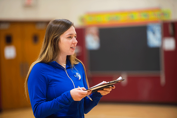 A woman with dark brown hair and blonde highlights stands in a gymnasium and holds a clipboard. She looks off into the distance. She is wearing a long-sleeved, blue ISU quarter-zip. Doors, a maroon-colored wall, and a large black bulletin board with a colorful border on the top are blurred but visible in the background.