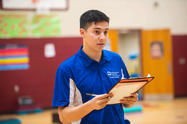 A white male student with dark hair stands in a gymnasium and holds a clipboard and pen. He wears a blue and white Indiana State polo shirt. He looks as if he is asking someone questions. The bottom of a basketball backboard is visible in the background, as is an open door, a maroon-colored wall, and some posters or signage.