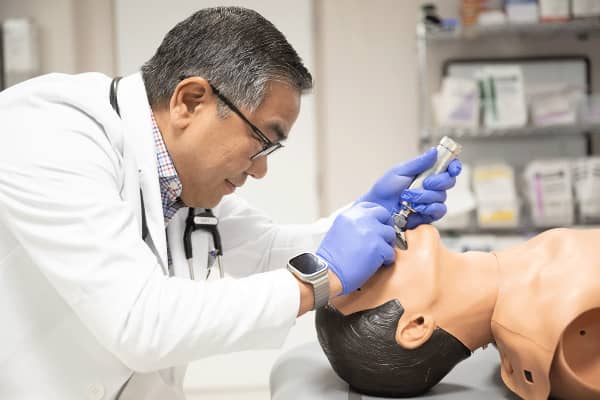 Middle aged male with white coat using a medical instrument to look down the throat of a medical practice mannequin.