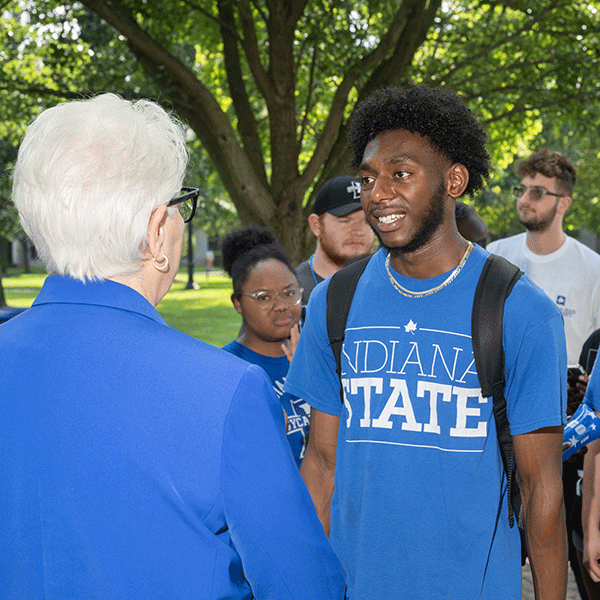 A black student wearing a blue Indiana State shirt standing outdoors talking to a woman with white hair and blue shirt.