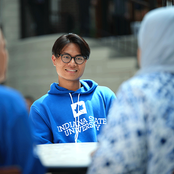 A student with glasses with a slight smile sitting at a table and looking towards a group of other students in the foreground with their backs to the camera. 