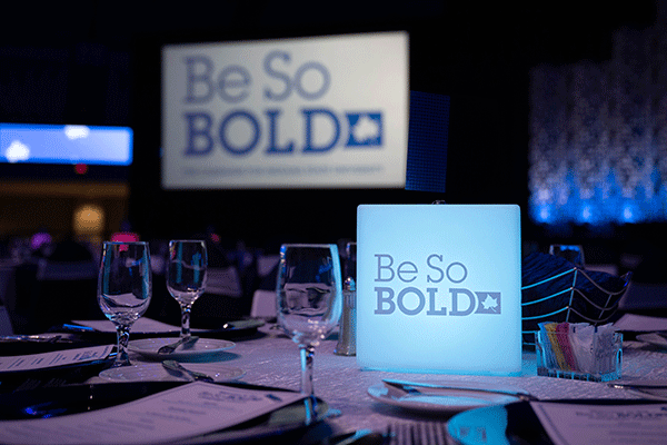 Circular table setting for a dinner in a dark room with a square illuminated center piece proclaiming “Be So Bold” with a large video screen in the background also showing “Be So Bold.”