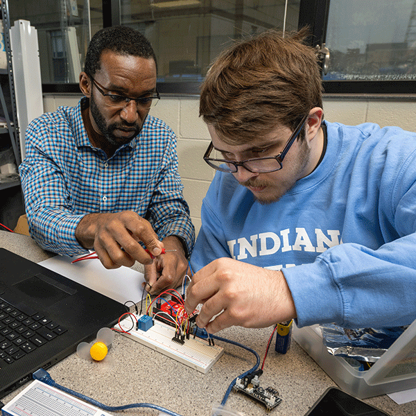 A Black male professor assisting a White student with brown hear working with wires on a board in a technology lab.