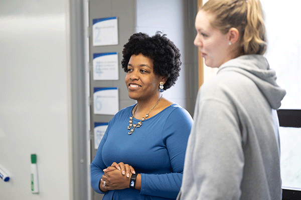 A Black professor speaking with a blond student with both looking from right to left and observing floor plans and drawings.