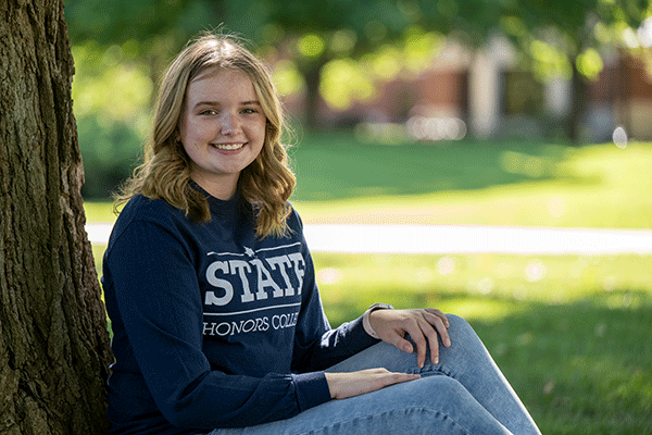 A female student with curly blonde hair sits with her back against a tree. She wears a dark blue STATE Honors College long-sleeved shirt and blue jeans. She is smiling and looking at the camera.