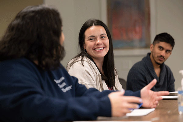 Three students sit at a desk in a classroom. On the right is a male student with black hair, wearing a grey sweater. He is looking at the other students. In the center is a female student with long brown hair. She wears a tan jacket and is smiling at the student on the left with short brown hair, wearing a blue sweatshirt. A painting is on the wall behind them.