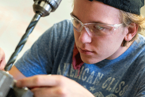 A white male student with blonde hair and wearing a black cap, glasses, and a powder-blue American Classic t-shirt, works on something that is out of focus at bottom left of frame. A drill is visible with a lengthy drill bit that extends down toward whatever he is holding in his fingers. The student’s bright blue eyes are visible as he focuses on his work.  