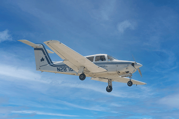 A white airplane with blue striping and blue lettering that says Indiana State University is pictured in flight against a blue sky with white clouds. N2878 can be made out on the side behind the wing. The three-wheel landing gear is down and the propeller appears to be in motion. A figure is faintly visible in the co-pilot’s seat. 