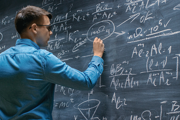 A man in a blue, long sleeved, collared shirt, who has short brown hair and wears glasses, writes equations on a chalk board that is full of them. 