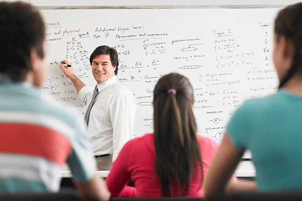 A white middle-aged male professor with short dark brown hair is standing inside a classroom in front of a whiteboard with mathematical equations written on the board in black. A black dry erase marker is visible in his right hand as he points to the board. He wears a white dress shirt, grey dress pants, and a grey necktie. He is smiling as he looks at the students sitting in the classroom. Their backs are to the camera. There are three students.