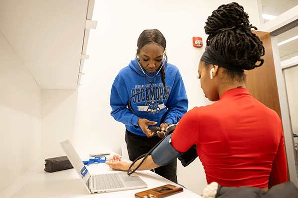 Two Black women pictured in a white exam room, with one measuring the other’s blood pressure. The woman taking the blood pressure wears black pants and a blue Sycamores sweatshirt with Sycamore Sam on the front. She has a stethoscope in her ears and is holding the pump to a blood pressure cuff in her hands. The woman wearing the blood pressure cuff is seated with her back to the camera. She wears a red top, a black bandanna, has a white earpiece in her ear, and her dreadlocked hair is piled atop her head.