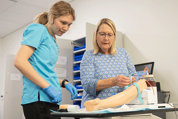  a White blond female student checks the blood pressure of a patient with back turned to the camera. 
