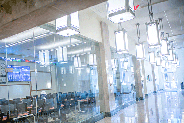 brightly lit hallway of a building with a a classroom/lab on the left side of the frame with glass doors and windows allowing the viewer to see a trading ticker and large screens I the room.