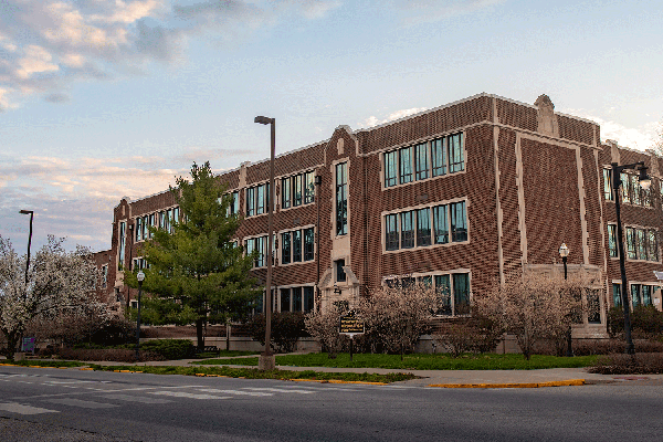 Exterior of a brick, three story building with large windows and trees in front with a blue sky and clouds and the intersection of two roads in front of the building