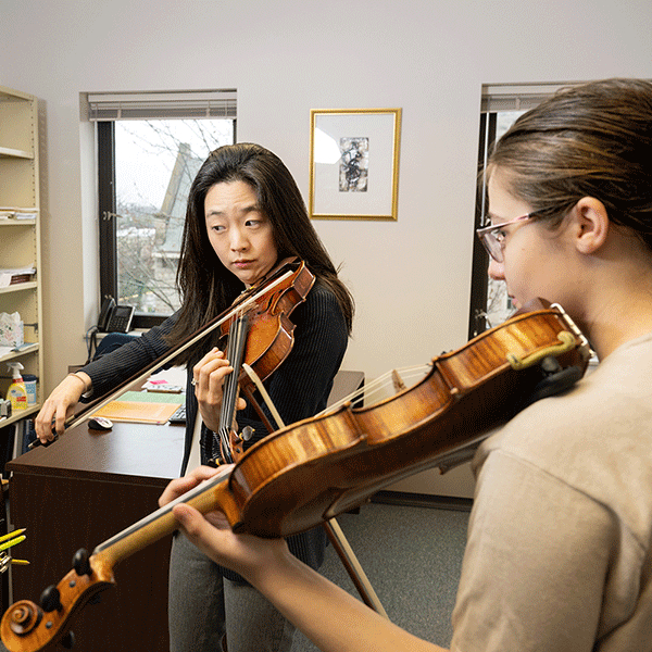 An Asian female professor with black shirt plays a violin while looking at sheet music as a White female student follows along.