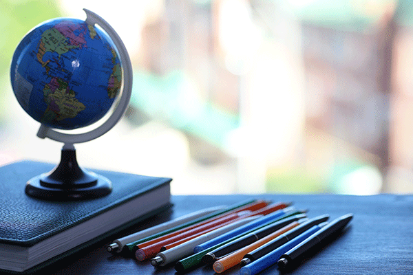 A globe sits on top of a book on a desktop with colored pencils beside the book