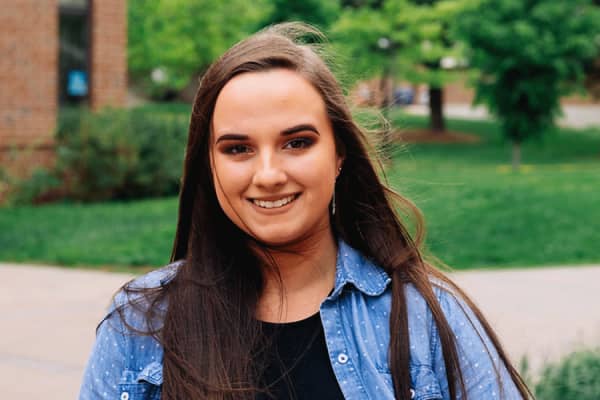 A white female student with long brown hair poses outside. She wears a blue jean jacket with a black undershirt. A brick building, a sidewalk, and a lawn with green trees are blurred in the background.