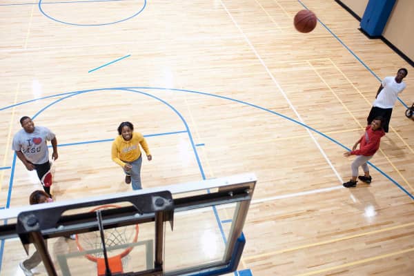 A group of five Black individuals playing basketball on a basketball court. The photo is taken from above the basketball hoop. The players wear a variety of white, red, yellow, grey, and black informal athletic gear (shorts, sweats, T-shirts, etc.). The basketball is visible mid-air, going towards the basketball hoop. The other players are looking up at the hoop.