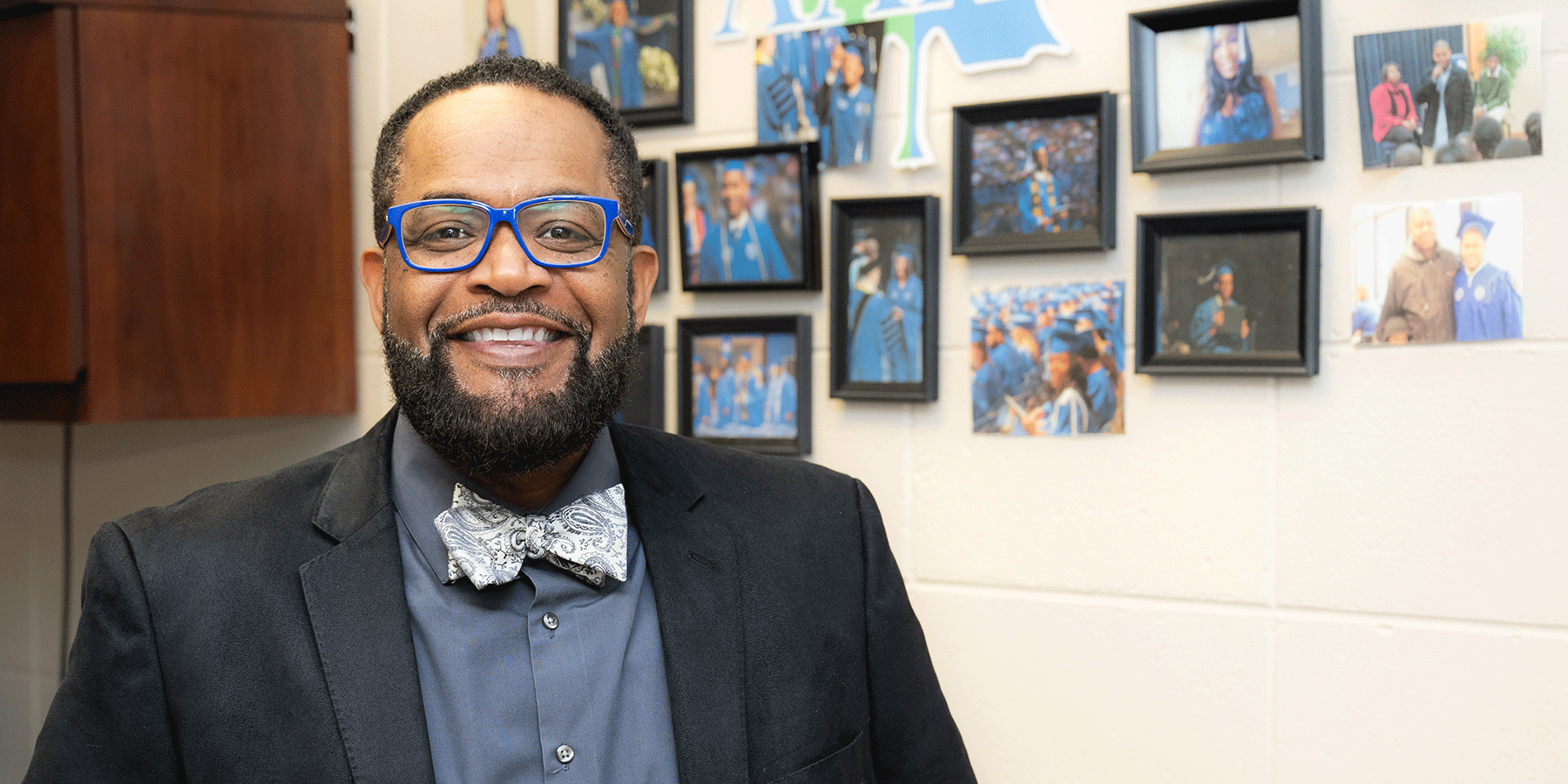 Black male with glasses and a suit posing in front of an office wall with pictures from a commencement ceremony with various students in caps and gowns in the background.