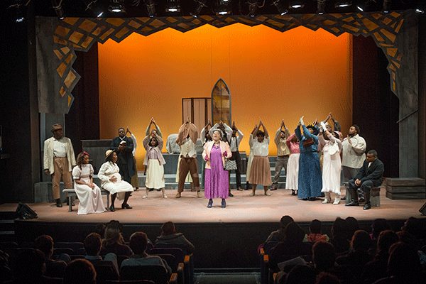 A theater performance on a stage. The cast includes 20+ Black performers wearing a variety of early 20th-century clothing. The stage is decorated with a dark orange stained-glass window in the back, a wardrobe changing divider, benches, and other orange stained-glass hanging from the ceiling. Several actors are visible with their arms raised in prayer. An audience sits in seats in the theater. 