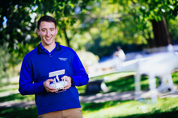 A white male student with short brown hair poses outside, wearing khaki pants and a blue zipped-up jacket with “Indiana State University” in white lettering in the top right corner of the jacket. He holds a white drone remote controller with a screen. A white drone is flying away from him, blurred in the photo. Green trees are visible in the background, also blurred in the photo. 