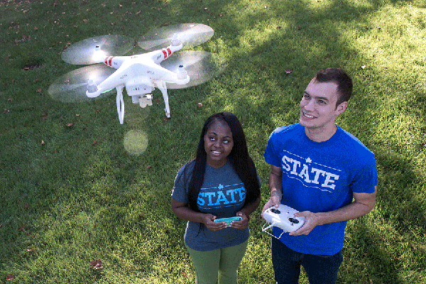 Two students are standing outside on a grassy lawn. On the left is a Black female student with long black hair. She wears green pants, red shoes, and a blue T-shirt with “STATE” in light blue lettering on the front. A blue smartphone is visible in her hands. On the right is a white male student with short brown hair. He wears denim jeans, black shoes, and a blue T-shirt with “STATE” in white lettering on the front. He holds a white drone remote controller while a white drone is flying above the students.