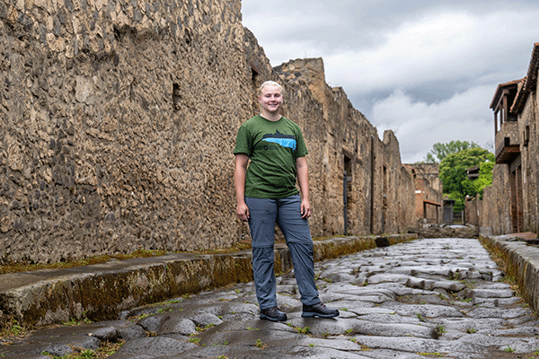 A female student standing on a rock road and looking at the camera while posing for a photo in the streets of the ancient city of Pompeii in Italy. Stone ruins of building are on each side of the roadway.