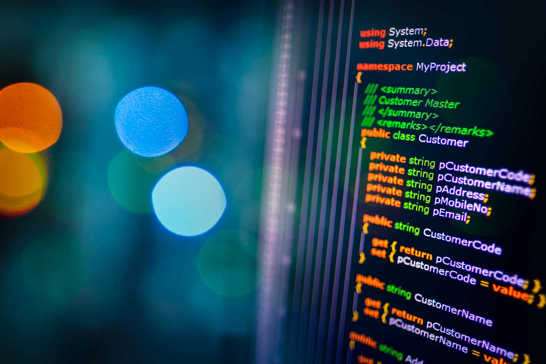 A computer monitor, shown at an angle on the right side of the screen, displays lines of code and words and phrases such as “Using System.Data,” “private string p.customer code,” “public string CustomerName,” etc. The words and codes appear in orange, green, and white digital text. Visible to the left half of the screen are large, out-of-focus dots of blue, green, and orange light.  