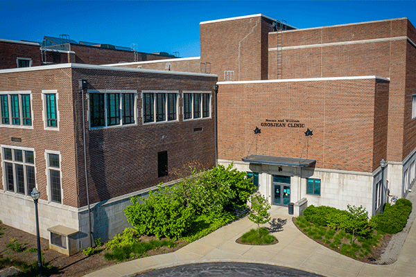 aerial image of a red-and-white brick, multi-story building that houses the Norma and William Grosjean Clinic. The clinc’s name is visible above the door. There is blue sky visible above and green grass and trees visible below. 