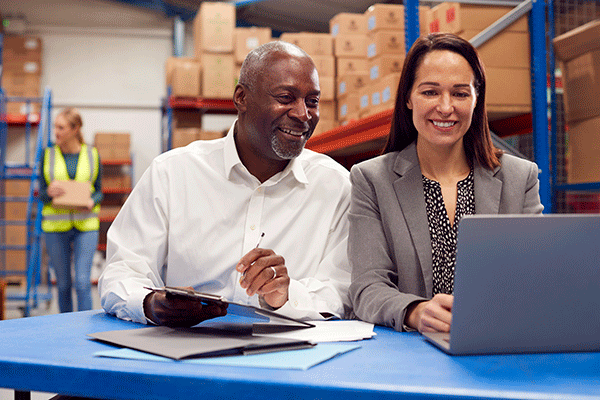 Two individuals are sitting at a blue table in a storage room with numerous brown packaging boxes stacked on shelves in the background. On the left is a Black male with short grey hair and matching facial hair. He wears a white long-sleeved dress shirt and a silver ring on his left hand. He holds a pen in his left hand and a clipboard in his right hand. He smiles as he looks at a grey laptop on the table. On the right is a White female with long, straight brown hair. She wears a grey blazer.