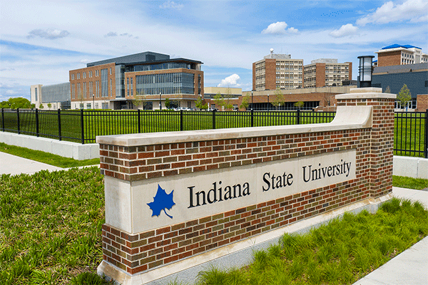 A large brick building with large windows is visible in the background of the image on the left, with three campus towers in the distance on the far right of the frame. In the foreground is a brick campus entryway surrounded by green grass, with a blue Sycamore leaf logo and “Indiana State University” on the sign.