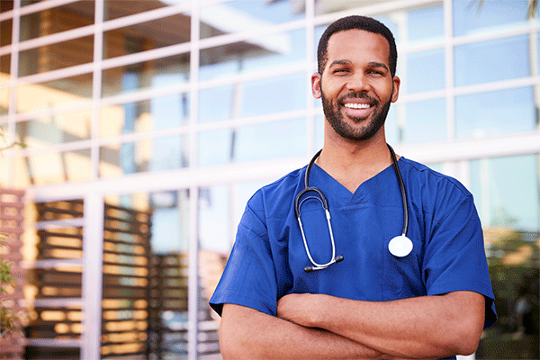 Black male with short hair, mustache and beard in blue nursing scrubs with arms crossed standing outside of a building with glass windows in the background. 