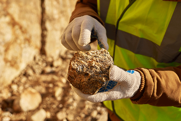Detail image of a person wearing a yellow safety vest with grey gloves holding a softball-sized rock. 