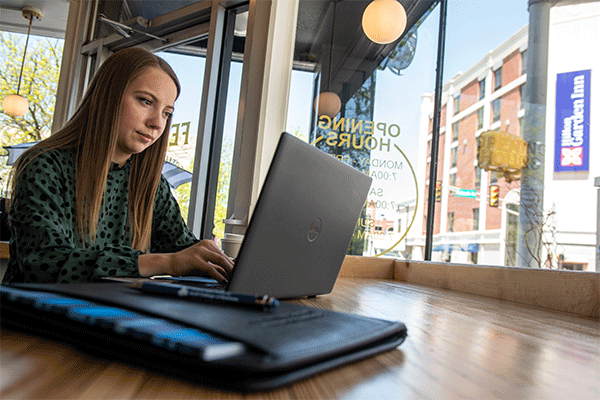 Female student with long brown hair wearing a dark green shirt sitting at a wooden table and working on a laptop in a coffee shop facing large windows looking out onto the street.  